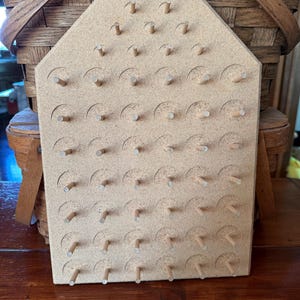 May include: A light brown, house-shaped wooden pegboard featuring a grid of circular indentations and protruding wooden pegs. The board has a flat base and an angled top, with a structured arrangement of pegs designed for organizing or displaying items.