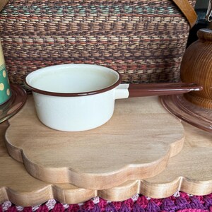 May include: A small, white enamel saucepan with a brown handle and rim sits on a wooden scalloped-edge tray. A woven picnic basket is in the background, along with a wooden jar and a pink glass plate.