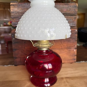 May include: A vintage oil lamp with a ruby red glass base and a white hobnail glass shade. The lamp has brass accents and a classic design. The lamp is a decorative item.