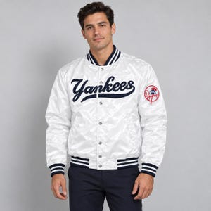 Puede incluir: Una chaqueta bomber de satén blanco con el texto Yankees en azul marino en el pecho, cuello y puños a rayas azul marino y blanco, botones de presión plateados y un parche circular con el logotipo de los Yankees en la manga izquierda.