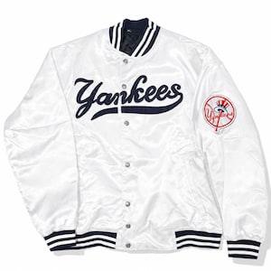 Può includere: Giacca bianca satinata dei Yankees con dettagli blu navy. Presenta la scritta "Yankees" in corsivo sul petto e una toppa con il logo della squadra sulla manica. La giacca ha una chiusura con bottoni e polsini e colletto a righe.