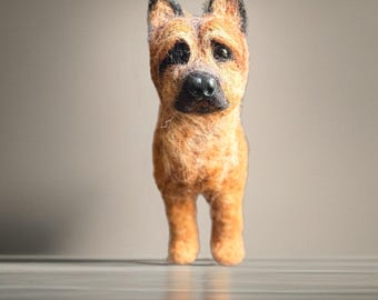 May include: A handcrafted felted German Shepherd dog figurine. The dog has a tan and black coat, with a detailed face and upright ears. The figurine is standing on a reflective surface, with a neutral background.