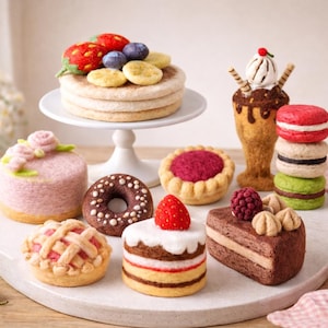Può includere: Una collezione di dessert in feltro in miniatura, tra cui una pila di pancake con frutta, una torta rosa con accenti floreali, una ciambella al cioccolato, una torta, una coppa gelato e macarons. I dessert sono disposti su un piatto bianco.