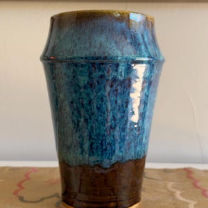 May include: A ceramic tumbler featuring a tapered silhouette with a glossy glaze. The upper portion displays a mottled blue and teal finish, while the lower section is a solid dark brown. The rim is smooth and rounded, showcasing a distinct transition between the two colored glaze sections.