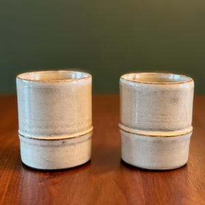 May include: Two cylindrical ceramic cups with a cream-colored glaze and a distinct horizontal ridge around the middle. The cups feature a smooth finish with subtle brown speckling and darker brown accents along the rim and the central ridge.