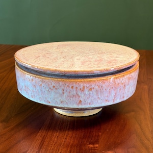 May include: A round ceramic lidded container featuring a speckled glaze in shades of soft pink, cream, and light blue. The vessel has a wide, cylindrical body with a slightly tapered base and a flat, circular lid that rests on the top rim.