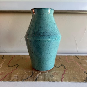 May include: A turquoise ceramic vase with a speckled finish and a distinct angular shoulder. The vessel features a glossy glaze, a narrow neck, and a wider body that tapers toward the base, showcasing a smooth, uniform texture throughout the piece.