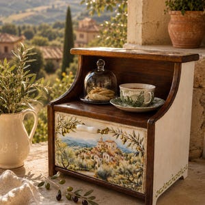 Puede incluir: Una panera de madera con un paisaje pintado de un pueblo en la ladera en el panel frontal del cajón. La caja tiene una superficie superior de madera oscura que sostiene una campana de cristal con galletas y una taza de cerámica.