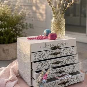 May include: A white wooden chest with four drawers, each featuring a white lace-patterned overlay and a small bronze-toned handle. The top drawer is inscribed with the text Le Petit Gateau. The chest is filled with colorful embroidery thread.