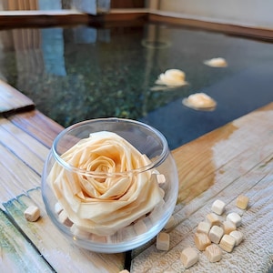 May include: A clear glass bowl with a large, light peach-colored rose inside, surrounded by small wooden cubes. The bowl sits on a wooden surface next to a Japanese-style bath with floating flower petals.