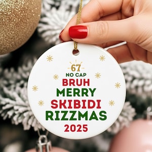 May include: A white ceramic Christmas ornament with gold accents and a gold string. The ornament features the phrases "-67- NO CAP BRUH MERRY SKIBIDI RIZZMAS 2025" in red and green text, with snowflake designs.