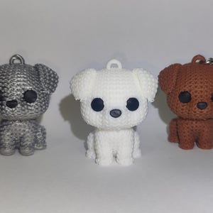 Pode incluir: Três pequenas figuras de cães em prata, branco e castanho. Cada cão tem olhos grandes e pretos e uma argola ou porta-chaves no topo. Os cães estão sentados, com as patas dianteiras visíveis.
