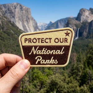 May include: A trapezoidal embroidered patch with a cream-colored top section featuring the text PROTECT OUR and a small star, and a dark brown bottom section with the text National Parks in a cursive font, all bordered by a cream-colored edge.