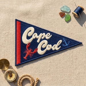 May include: A triangular navy blue felt pennant featuring the embroidered white text Cape Cod, a red lobster, and a small blue anchor. The pennant has a red vertical stripe on the left side and is displayed with a compass, thread, and sea glass.