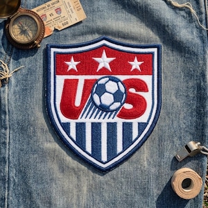 May include: A shield-shaped embroidered patch featuring the red, white, and blue US soccer logo with three stars, the letters U and S, and a soccer ball graphic. The patch is displayed on blue denim fabric alongside a vintage compass and a ticket.