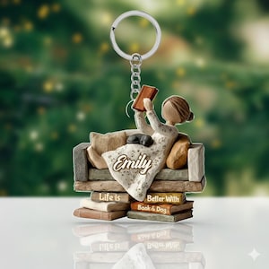 May include: A keychain featuring a woman reading a book while reclining on a couch, with a small dog. The keychain has a silver ring and chain. The couch is atop a stack of books with the text "Life is Better With Book & Dog" and the name "Emily".
