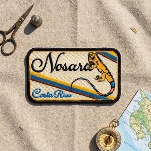 May include: A rectangular embroidered patch featuring the text Nosara and Costa Rica in black and blue script. The design includes a yellow iguana with a blue and pink tail, set against a cream background with diagonal blue and brown stripes.