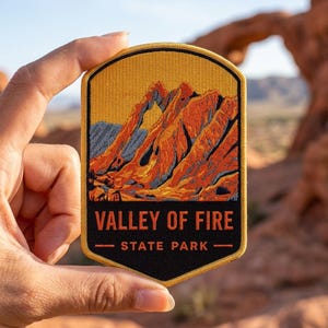 May include: A shield-shaped embroidered patch featuring a landscape of orange and red rock formations against a blue sky. The bottom section is black with the orange text VALLEY OF FIRE STATE PARK, framed by a yellow border and stitching.