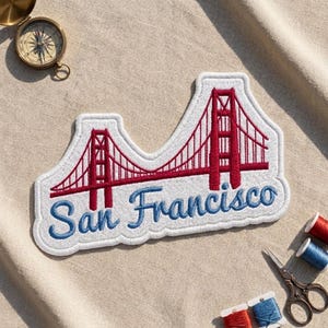 May include: An embroidered patch featuring a red Golden Gate Bridge design on a white background with the blue text San Francisco underneath. The patch has a white border and is positioned near a brass compass and sewing supplies.