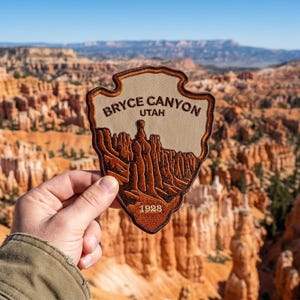 May include: A shield-shaped embroidered patch featuring a tan background with dark brown stitching. The text reads BRYCE CANYON UTAH at the top and 1928 at the bottom. A detailed dark brown landscape illustration of rock formations fills the center.