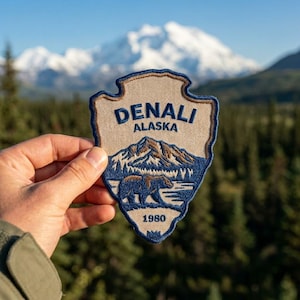 May include: A shield-shaped embroidered patch featuring a beige background with a dark blue border. The text reads DENALI ALASKA and 1980. The design includes a brown and blue mountain range above a brown bear silhouette near a body of water.