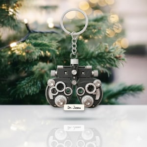 May include: A black and silver phoropter keychain with a silver chain and keyring. The keychain has multiple dials and lenses, and a small white tag that reads "Dr. Jenna". The background is a blurred Christmas tree.