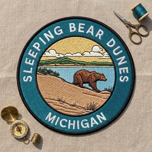 May include: A circular embroidered patch featuring a brown bear walking on a sandy dune with a blue lake and green hills in the background. The border is teal with the white text SLEEPING BEAR DUNES MICHIGAN. Includes scissors and a compass.