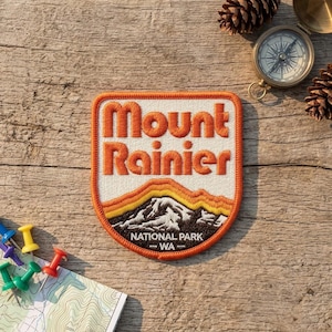 May include: A shield-shaped embroidered patch featuring the text Mount Rainier in orange thread on a white background. Below the text is a stylized mountain graphic in yellow, orange, and dark brown, with the text NATIONAL PARK WA at the bottom.