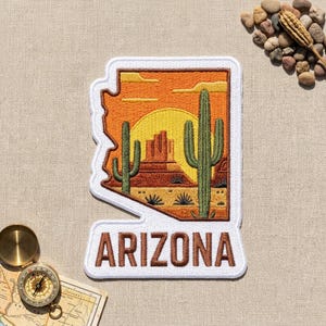May include: An embroidered patch shaped like the state of Arizona with a white border. The design features a desert landscape with a yellow sun, orange sky, brown rock formations, and green saguaro cacti. The word ARIZONA is stitched in brown.
