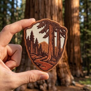 May include: A shield-shaped embroidered patch featuring a landscape scene with tall brown trees, a winding path, and a mountain in the background. The design uses shades of brown and cream thread against a light-colored background with a brown border.