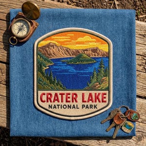 May include: An embroidered patch featuring a landscape of Crater Lake National Park with a blue lake, green trees, and orange sky, placed on blue denim fabric. A brass compass and a metal keyring with small charms are positioned nearby.