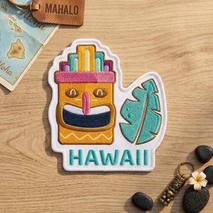 May include: An embroidered patch featuring a yellow tiki figure with a pink nose, a wide dark blue mouth, and a teal tropical leaf. The patch has a white border and the word HAWAII stitched in teal letters at the bottom on a white background.