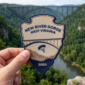 May include: A shield-shaped embroidered patch featuring a tan background with dark blue borders and text. The patch displays the words NEW RIVER GORGE WEST VIRGINIA, an illustration of an arched bridge, a small parachute icon, and the year 2020.