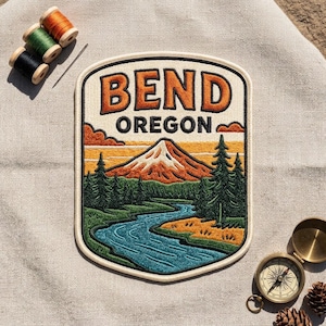 May include: An embroidered patch featuring the text BEND OREGON above a landscape of a snow-capped mountain, a winding blue river, and green pine trees. The patch has a cream border and is surrounded by three spools of thread and a brass compass.