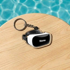May include: A VR headset keychain with a black and white design. The keychain has a silver ring and chain. The VR headset has the word "Name" printed on the front. The keychain is on a wooden surface.