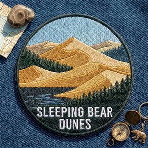 May include: A circular embroidered patch featuring a landscape of golden sand dunes, dark green pine forests, and a blue sky. The text SLEEPING BEAR DUNES is stitched in white at the bottom. The patch has a dark green border and is round in shape.