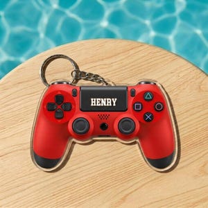 May include: Red video game controller keychain with the name "HENRY" printed on the center. The keychain has a silver ring and chain. The controller has black accents and the standard button layout.