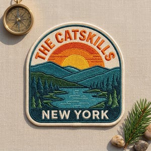 May include: An embroidered patch featuring a landscape of The Catskills, New York. The design shows a yellow and orange sun rising over teal mountains and a blue lake surrounded by green pine trees. The text reads THE CATSKILLS and NEW YORK.