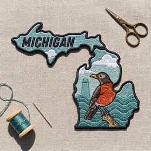 May include: An embroidered patch shaped like the state of Michigan with a black border. The patch features the word MICHIGAN in black text, a robin bird perched on a branch, stylized blue waves, a lighthouse, and white clouds on a light blue background.