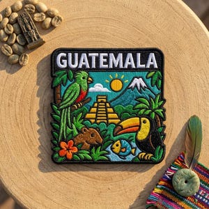 May include: A square embroidered patch featuring the word GUATEMALA in white text. The design includes a green quetzal bird, a yellow Mayan pyramid, a sun, a volcano, a toucan, a small fish, and a brown animal, all set against a blue sky.