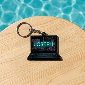 May include: A black laptop-shaped keychain with the name "JOSEPH" in neon blue on the screen. The keychain has a silver keyring and chain. The background is a light wood surface with a blue water reflection.
