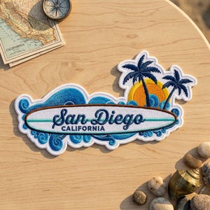 May include: An embroidered patch featuring a white surfboard with the text San Diego CALIFORNIA in dark blue script. The design includes blue ocean waves, a yellow sun, and two dark blue palm trees against a white background border.