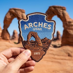 May include: An embroidered patch shaped like a National Park Service arrowhead, featuring a blue sky gradient, a brown landscape with the Delicate Arch rock formation, and the white text ARCHES UTAH 1971 with a dark brown stitched border.