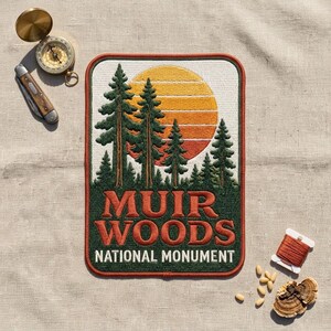 May include: A rectangular embroidered patch featuring a forest of green pine trees against a yellow and orange sunset. The text MUIR WOODS NATIONAL MONUMENT is stitched in orange and white. A brass compass, a pocket knife, thread, and fungi are nearby.