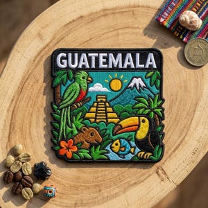 May include: A square embroidered patch featuring the word GUATEMALA in white text. The design includes a green quetzal bird, a yellow Mayan pyramid, a blue mountain, a toucan, a small fish, a brown animal, a bright orange flower, and a yellow sun.