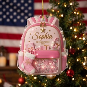 May include: A pink backpack-shaped Christmas ornament featuring the name Sophia, the text Kindergarten 2026, a small pink stocking, a gift box, a tiara, and a clear front pocket filled with pink and white star and heart-shaped confetti.