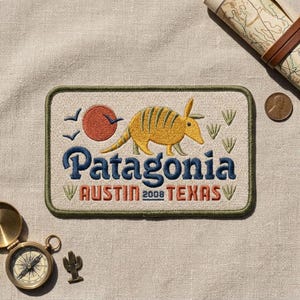 May include: A rectangular embroidered patch featuring a yellow armadillo, a red sun, and blue birds. The text reads Patagonia, Austin, 2008, Texas. The patch has a dark green border and is set against a light beige fabric background with a compass.