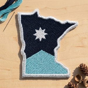 May include: An embroidered patch in the shape of the state of Minnesota. The patch features a dark navy blue upper section with a white eight-pointed star and a light blue lower section, all outlined with a thick white stitched border.