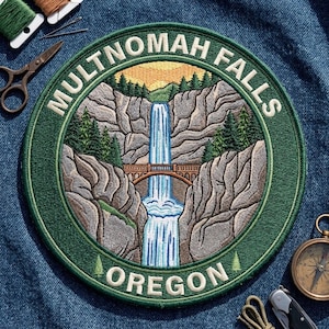 May include: A circular embroidered patch featuring a landscape of Multnomah Falls in Oregon. The design includes a waterfall cascading over rocky cliffs, a stone bridge, and evergreen trees under a yellow sky. The text reads Multnomah Falls Oregon.