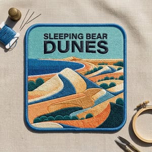 May include: A square embroidered patch featuring a landscape of Sleeping Bear Dunes with tan sand hills, blue water, and green vegetation. The text SLEEPING BEAR DUNES is stitched in dark blue at the top. The patch has a blue border and rounded corners.